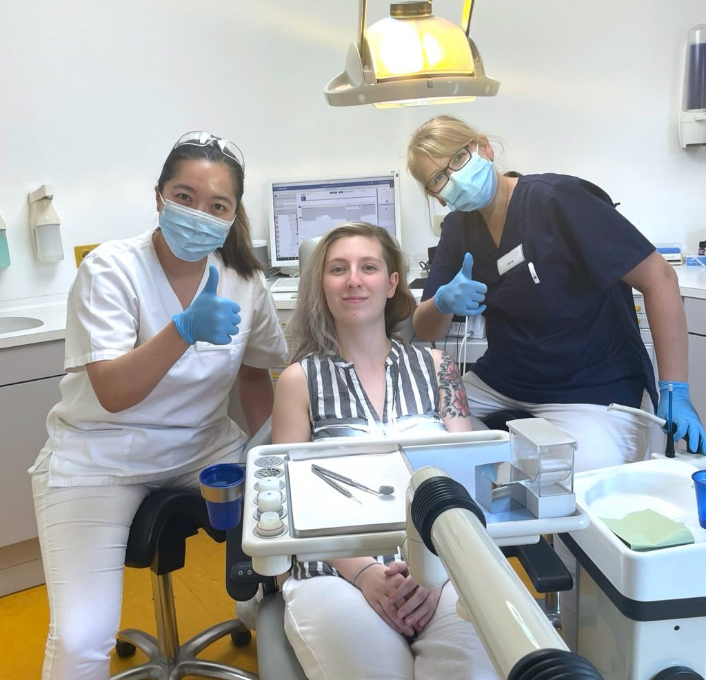 Links sitzt eine Zahnärztin, rechts sitzt eine Zahnmedizinische Fachangestellte. Sie behandeln die Patientin, die zwischen ihnen im Behandlungsstuhl liegt. Zahnärztin und Zahnmedizinische Fachangestellte zeigen ein Daumen hoch in die Kamera.
