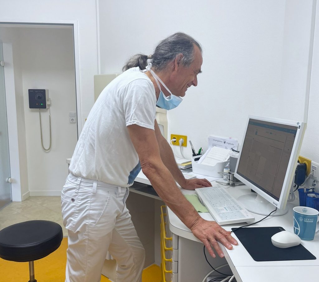 Ein Zahnarzt steht im Behandlungszimmer vor einem Computerbildschirm und schaut sich etwas auf dem Bildschirm an.