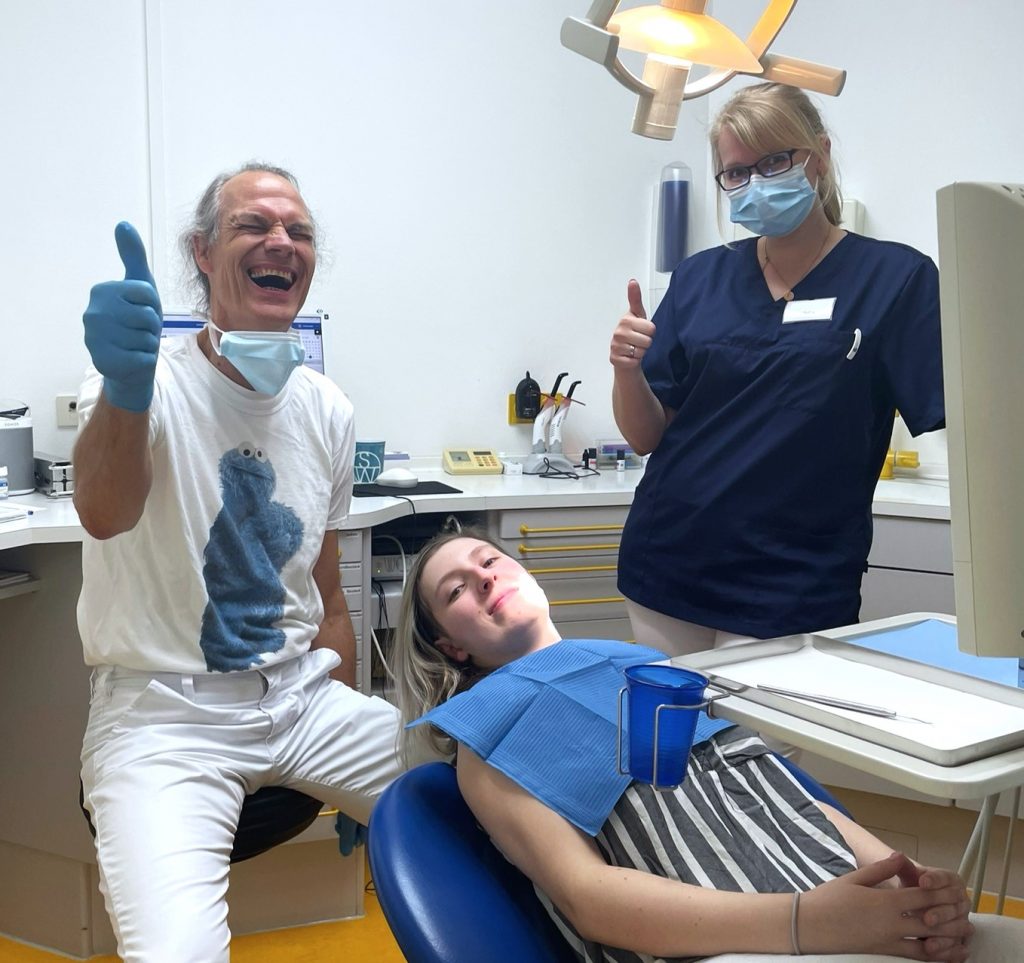 Links sitzt ein Zahnarzt, in der Mitte liegt eine Patientin in einem blauen Behandlungsstuhl, rechts steht eine zahnmedizinische Fachangestellte. Die zahnmedizinische Fachangestellte und der Zahnarzt zeigen ein Daumen hoch in die Kamera.