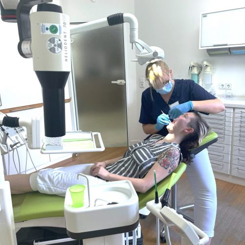 Im Vordergrund liegt eine Patientin in einem Behandlungsstuhl, im Hintergrund sitzt eine zahnmedizinische Fachangestellte, die die Patientin behandelt.