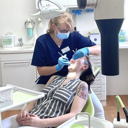 Im Vordergrund liegt eine Patientin in einem Behandlungsstuhl, im Hintergrund sitzt eine zahnmedizinische Fachangestellte, die die Patientin behandelt.