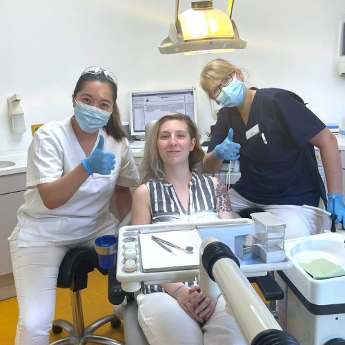 Links sitzt eine Zahnärztin, rechts sitzt eine Zahnmedizinische Fachangestellte. Sie behandeln die Patientin, die zwischen ihnen im Behandlungsstuhl liegt. Zahnärztin und Zahnmedizinische Fachangestellte zeigen ein Daumen hoch in die Kamera.