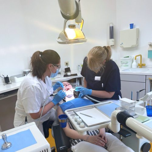 Links sitzt eine Zahnärztin, rechts sitzt eine Zahnmedizinische Fachangestellte. Sie behandeln die Patientin, die zwischen ihnen im Behandlungsstuhl liegt.